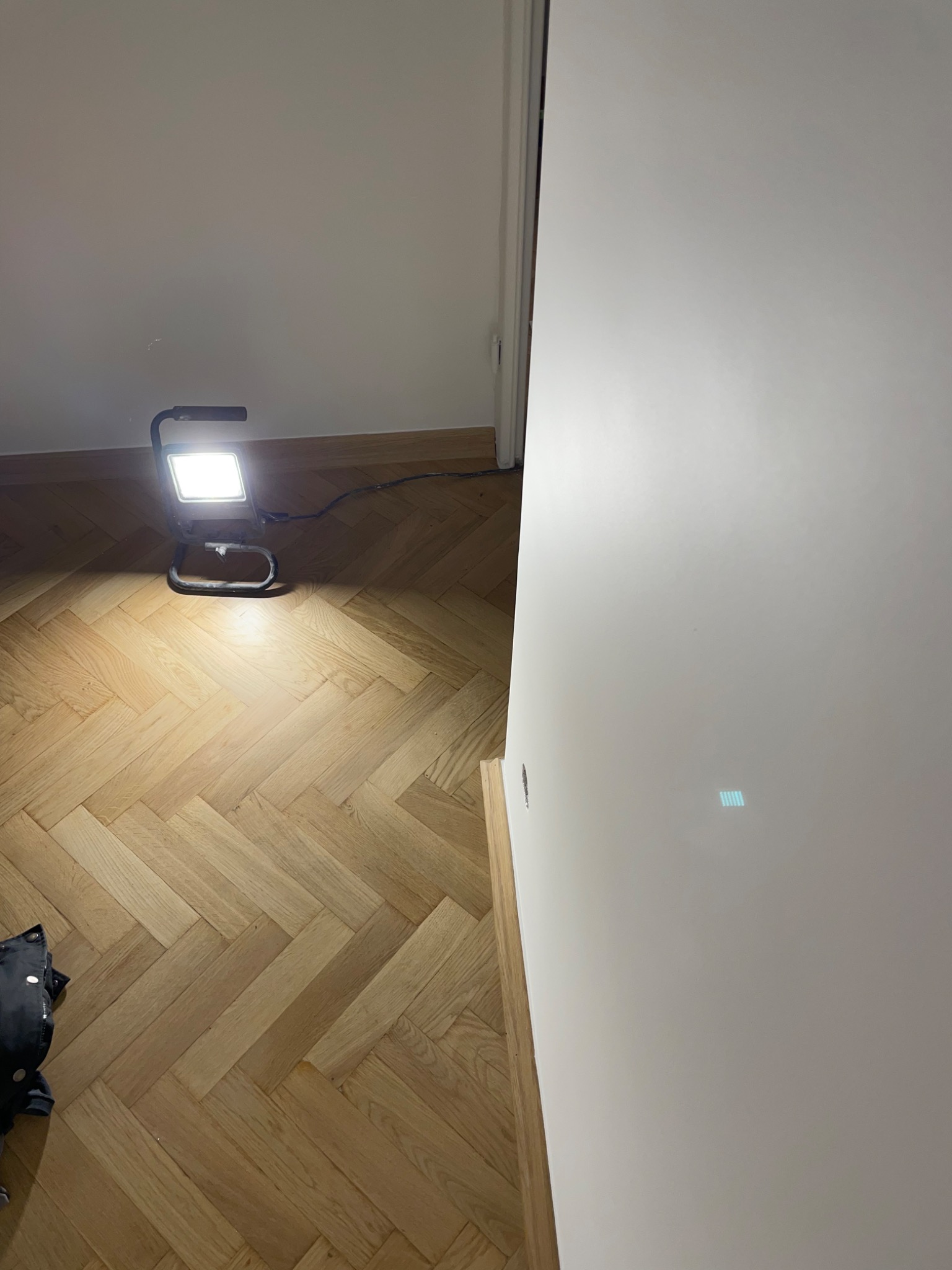 Świeżo pomalowana, biała ściana podświetlona lampą roboczą na tle parkietu ułożonego w jodełkę.