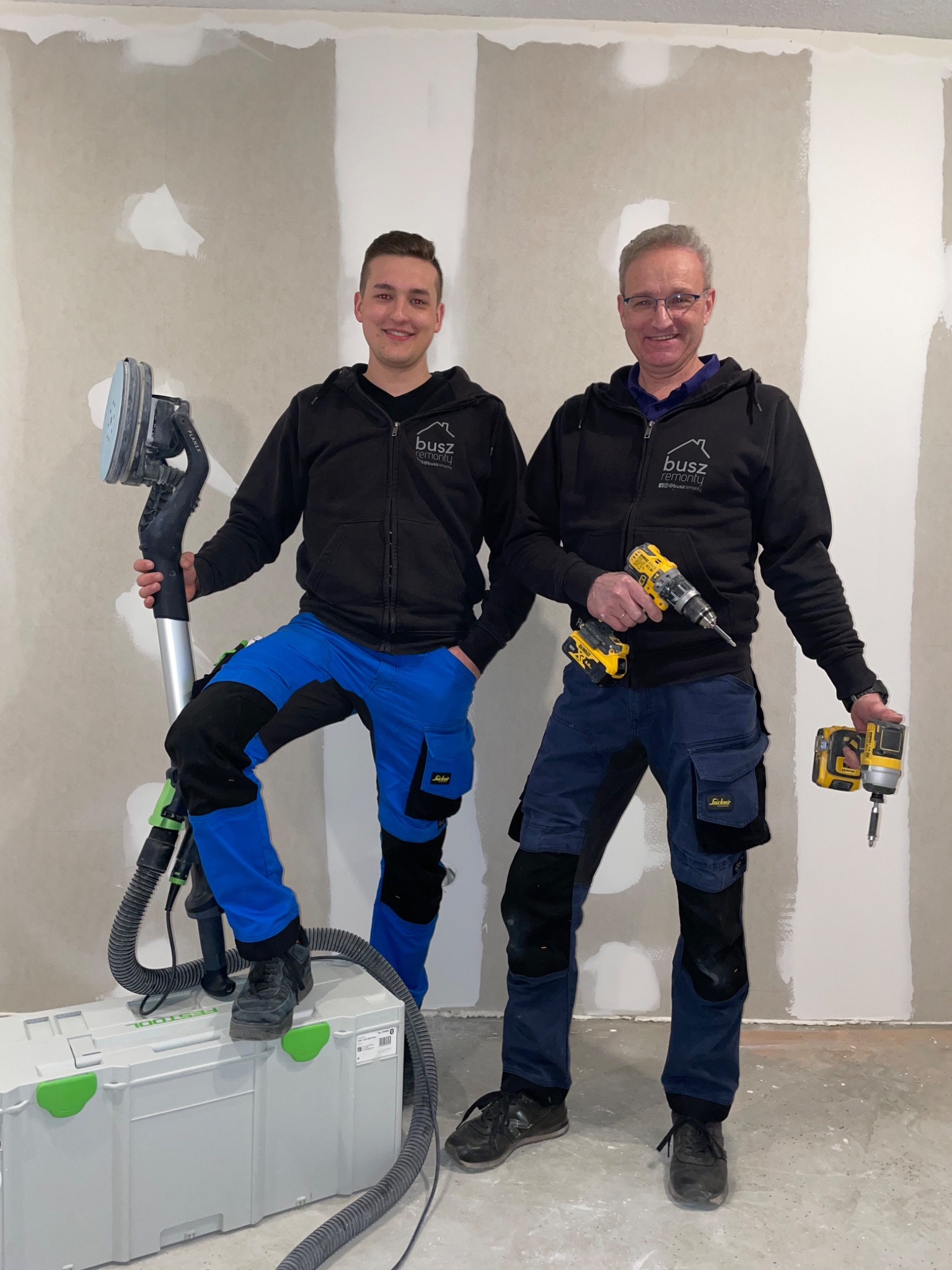 Dwóch mężczyzn w odzieży roboczej, prezentujących szlifierkę do gładzi i wkrętarkę na tle ściany po szpachlowaniu, z widocznymi łatami i nierównościami.