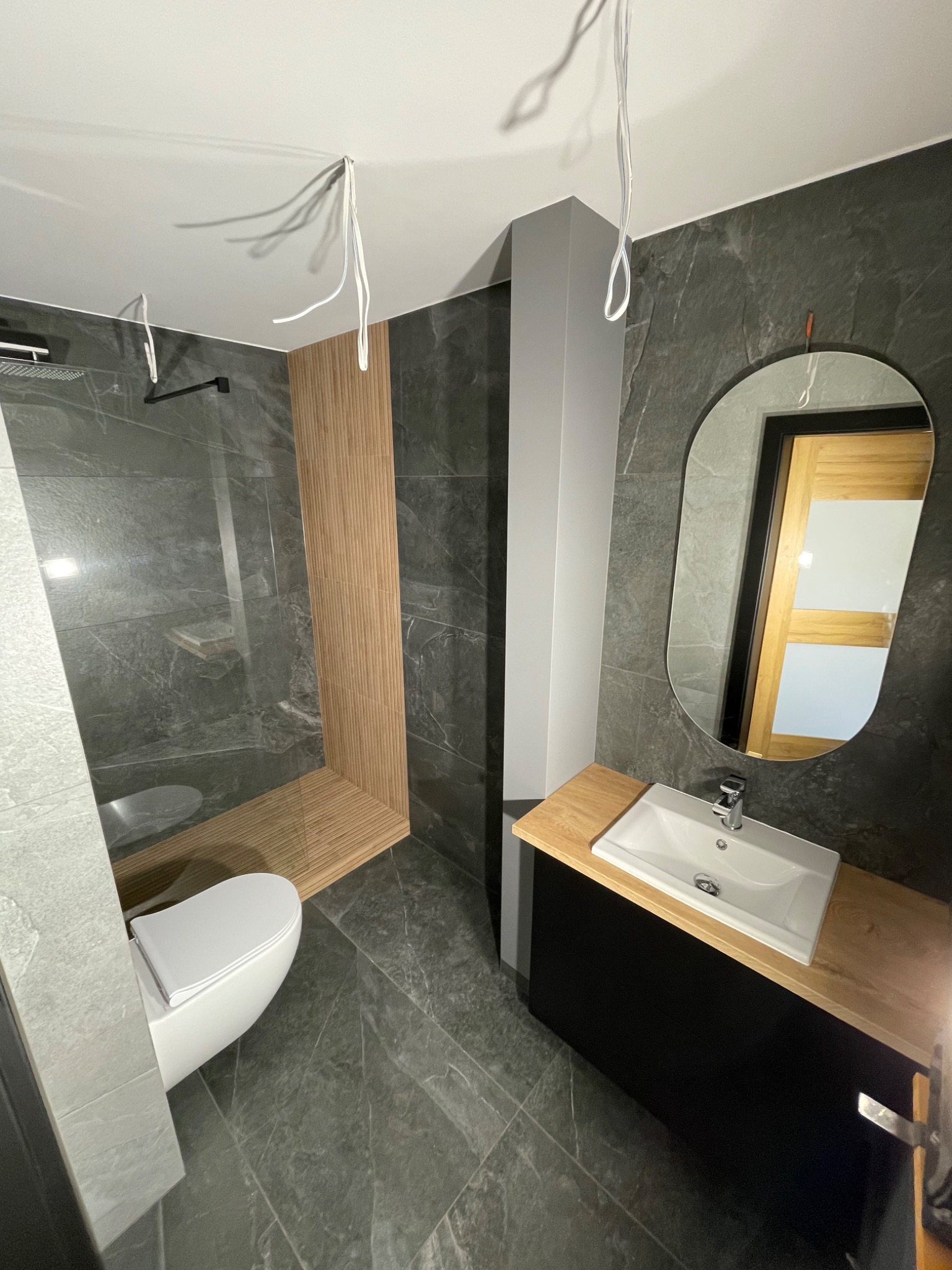 Nowoczesna łazienka z szarymi płytkami imitującymi marmur, kabiną prysznicową typu walk-in z drewnianymi akcentami, owalnym lustrem i umywalką nablatową na czarnej szafce z blatem z jasnego drewna;...