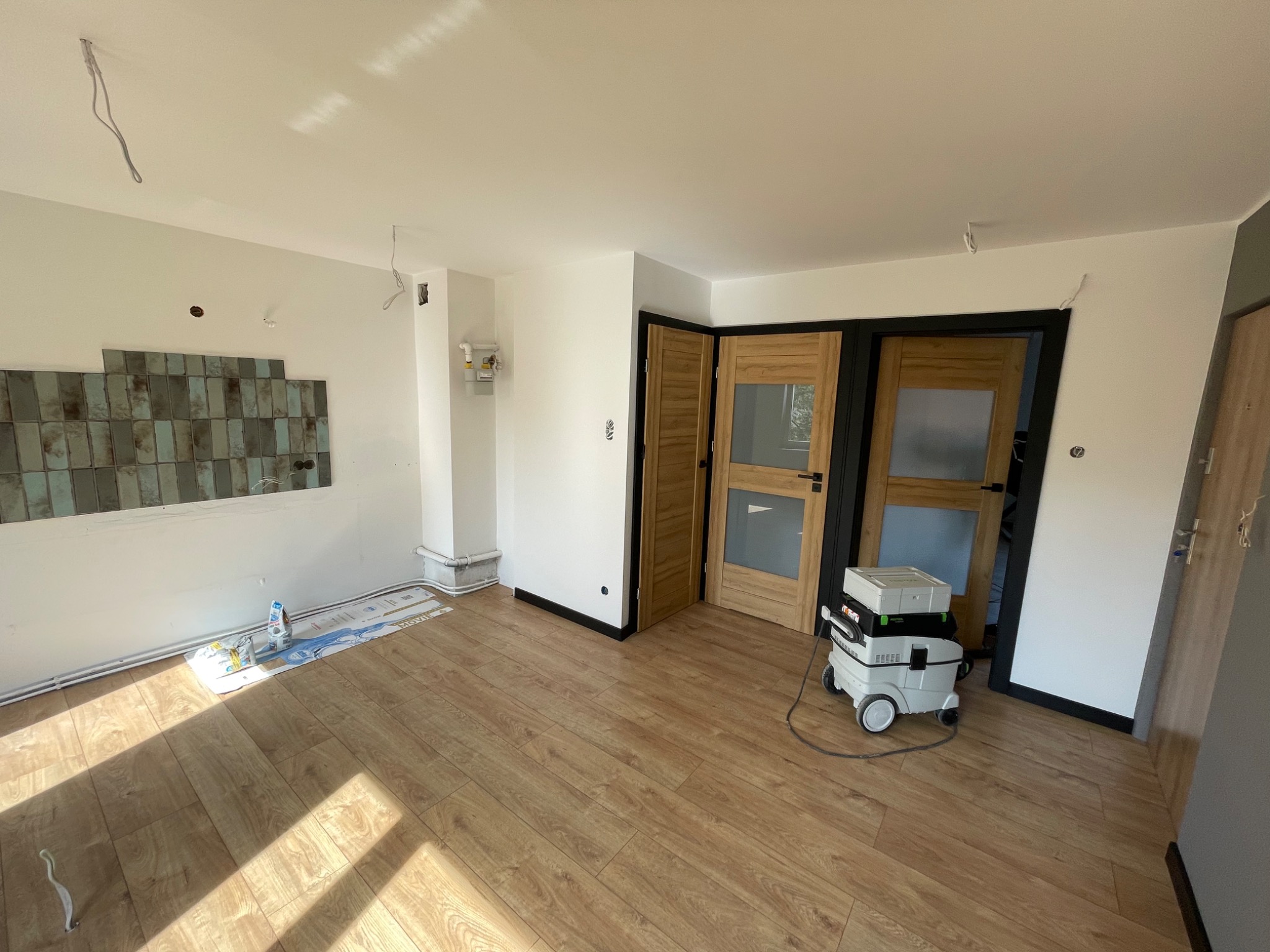 Pokój w trakcie prac wykończeniowych: położona podłoga, częściowo ułożona glazura na ścianie, świeżo pomalowane ściany i sufit, widoczny odkurzacz przemysłowy i materiały budowlane.