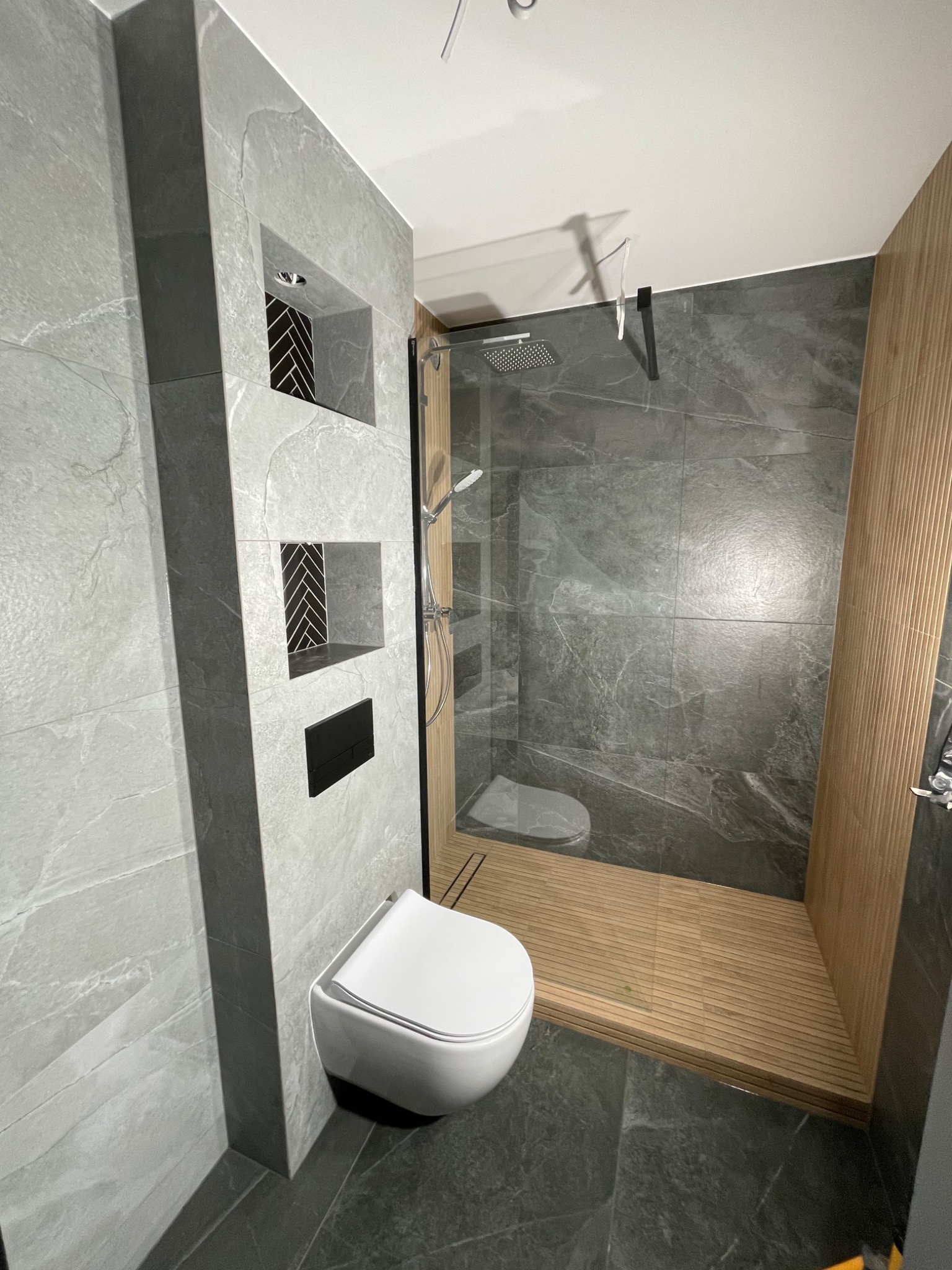 Nowoczesna łazienka z szarymi płytkami imitującymi marmur, drewnianym wykończeniem prysznica, podwieszaną toaletą i wnękami z czarno-białą mozaiką w ścianie.