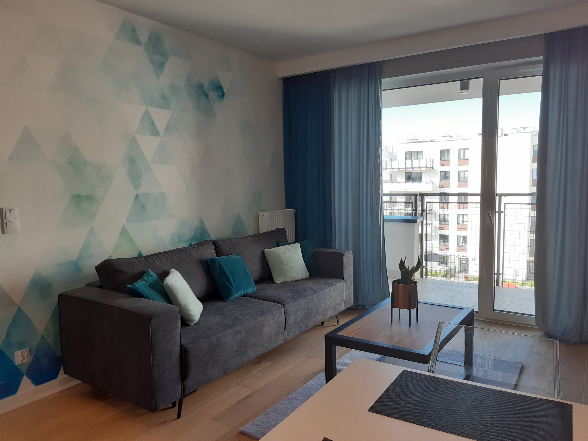 Nowoczesny salon z szarą sofą, turkusowymi poduszkami i geometryczną tapetą w odcieniach błękitu i zieleni. Stolik kawowy z rośliną w tle okna balkonowego z widokiem na budynek mieszkalny.