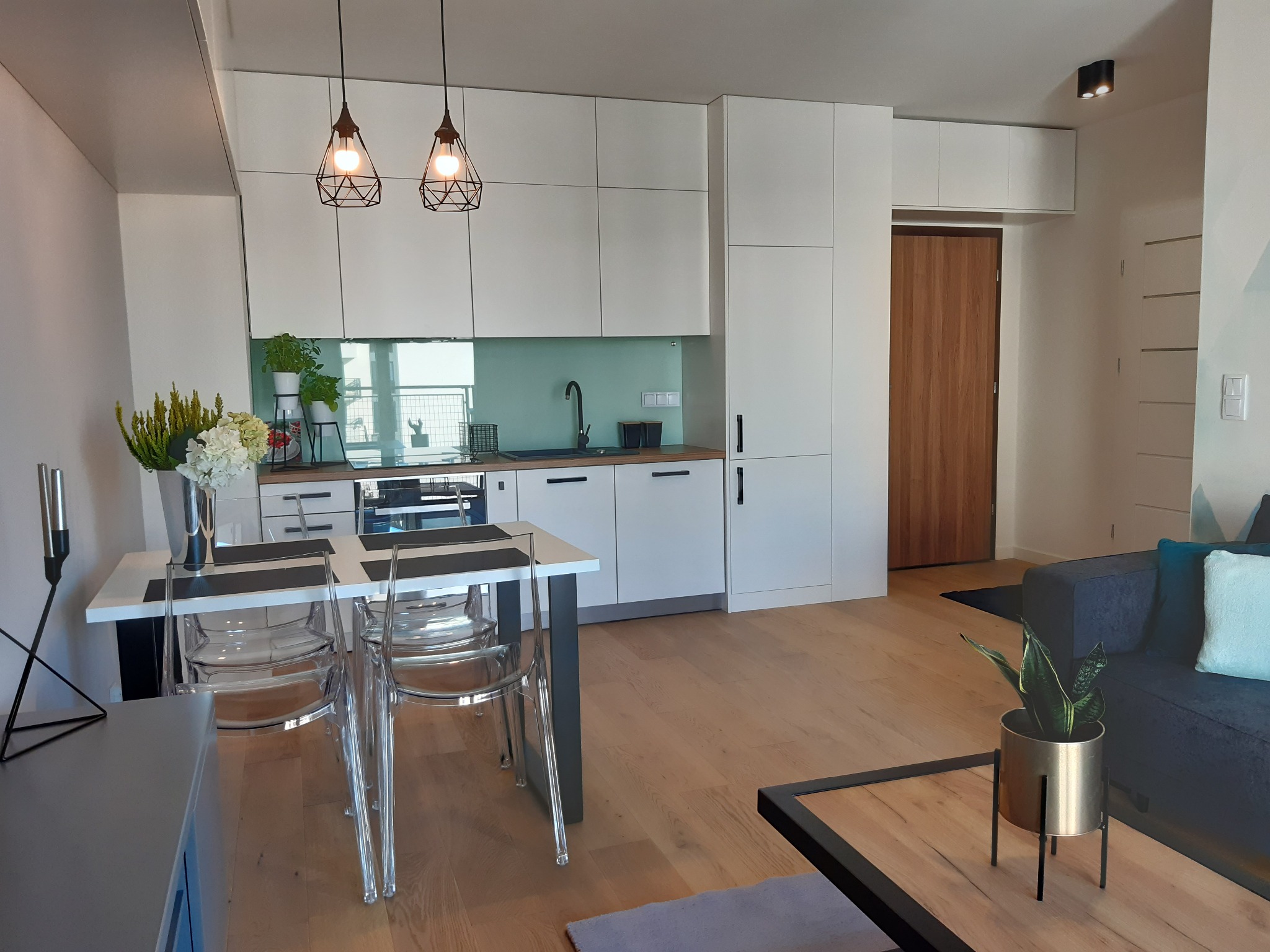 Nowoczesne wnętrze mieszkania z otwartą kuchnią, białymi szafkami, drewnianym blatem i podłogą, transparentnymi krzesłami przy stole i ozdobnymi lampami wiszącymi.
