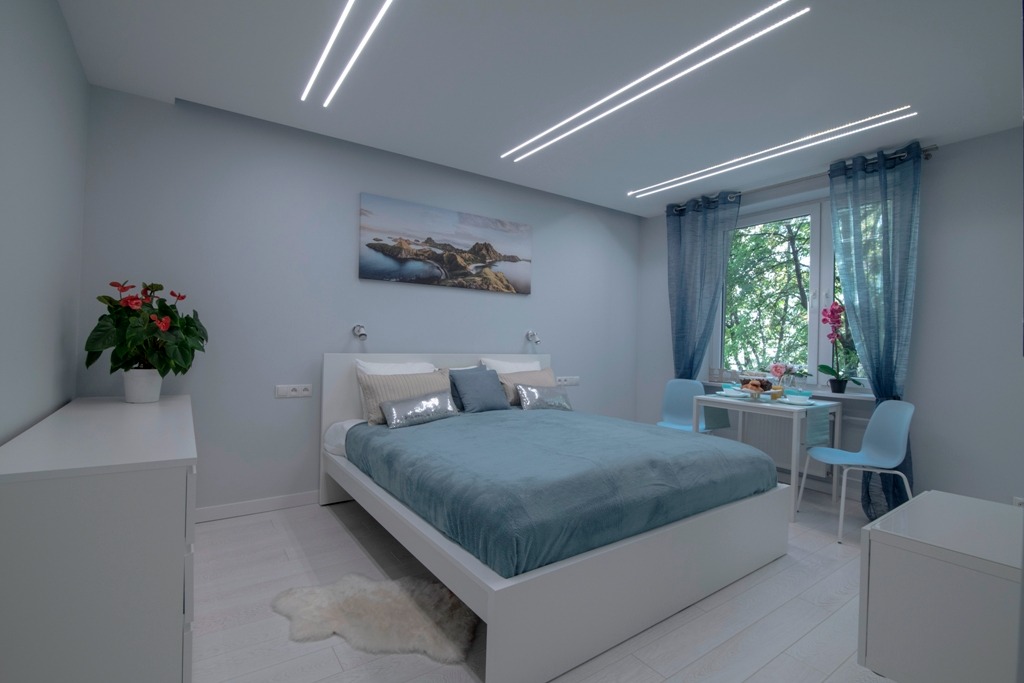 Jasna sypialnia w odcieniach błękitu z białymi meblami i dekoracyjnym oświetleniem LED na suficie, obraz na ścianie i stolik śniadaniowy przy oknie.