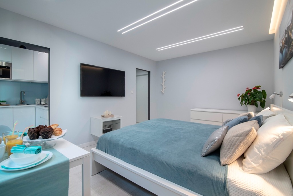 Nowoczesna aranżacja wnętrza mieszkania typu studio z łóżkiem, aneksem kuchennym, telewizorem na ścianie i śniadaniem na stoliku.