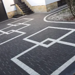 Czarna kostka brukowa z białymi, geometrycznymi wstawkami tworząca wzór na podjeździe przed domem z garażem i schodami.