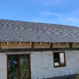 Surowy stan domu z białego betonu komórkowego w Grudziądzu, z zamontowanymi brązowymi oknami i pokrytym gontem dachem, widoczne drewniane elementy konstrukcyjne pod okapem.