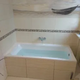 Wanna z obudową wykończoną płytkami imitującymi marmur, wypełniona wodą, z dekoracyjnym pasem płytek imitujących kamienie i górnym pasem z motywem plaży i łodzi, widoczny gumowy młotek przy...