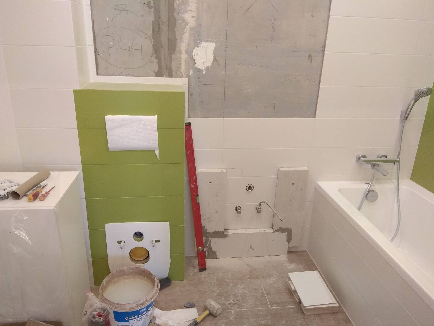 Niedokończona instalacja podtynkowa WC w łazience z częściowo położonymi białymi płytkami, widoczny stelaż w odcieniach zieleni, poziomica, wiadro z zaprawą i fragment wanny.