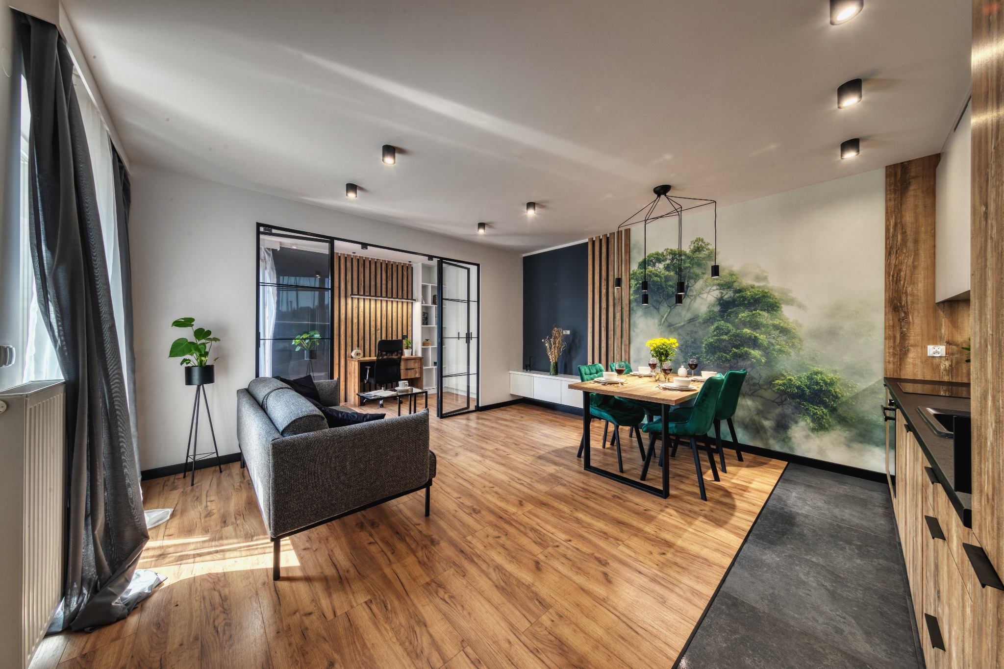 Nowoczesne wnętrze z drewnianą podłogą, szarą sofą i stołem jadalnym z zielonymi krzesłami, ozdobione fototapetą z motywem leśnym i drewnianymi lamelami.