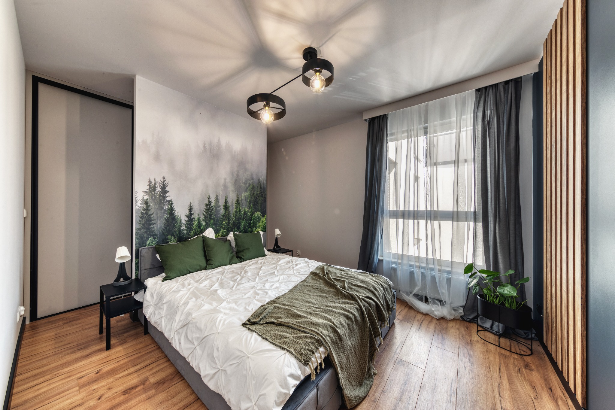 Nowoczesna sypialnia z fototapetą leśną, drewnianą podłogą i czarnymi akcentami: lampy sufitowe, stoliki nocne i donica z rośliną.