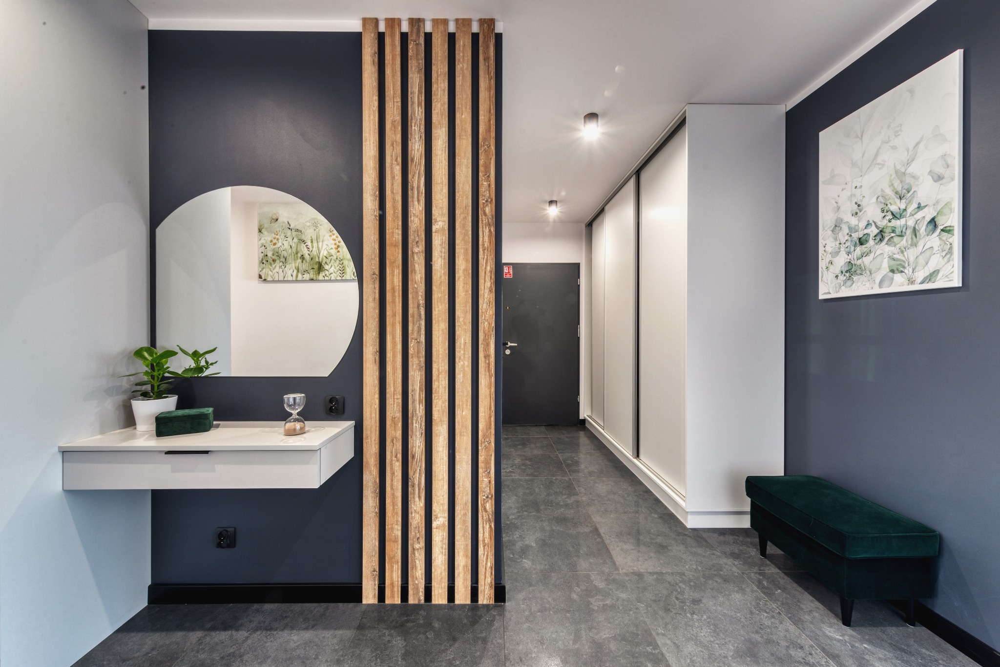 Nowoczesny przedpokój z szarą podłogą, granatowymi ścianami, okrągłym lustrem nad białą konsolą z kwiatem i klepsydrą, drewnianą ażurową ścianką działową oraz zieloną pufą.