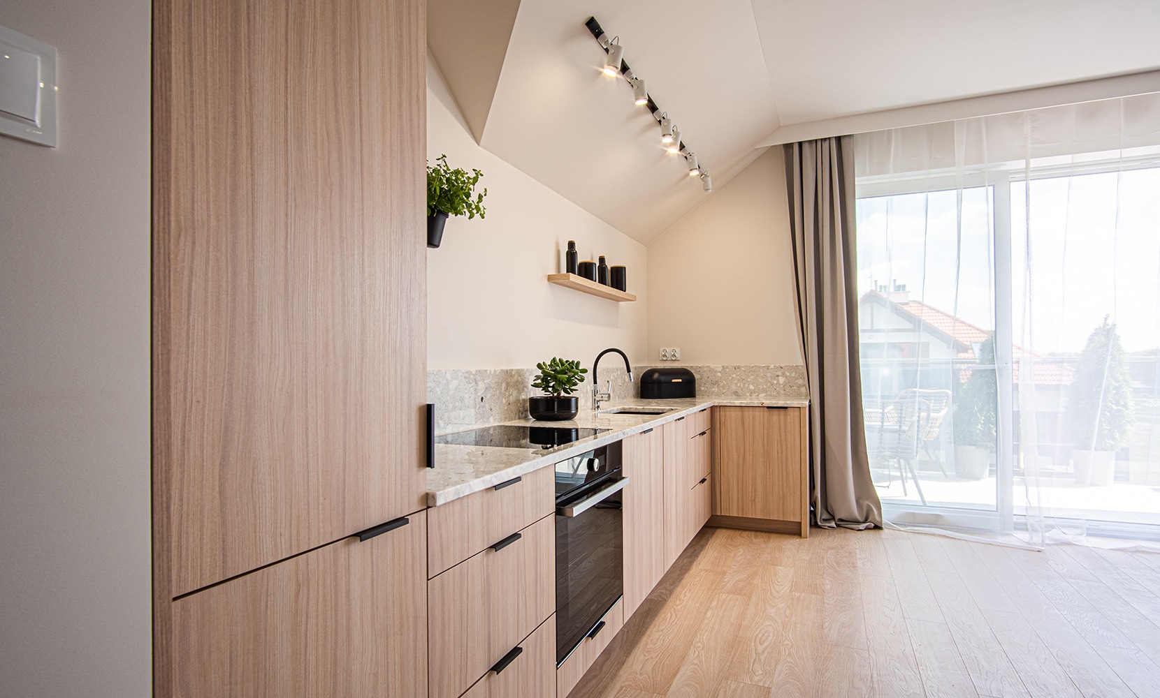 Jasna kuchnia w skandynawskim stylu z drewnianymi frontami szafek, blatem z jasnego kamienia, czarnymi uchwytami i oświetleniem szynowym na skośnym suficie, z widokiem na balkon przez duże okno.