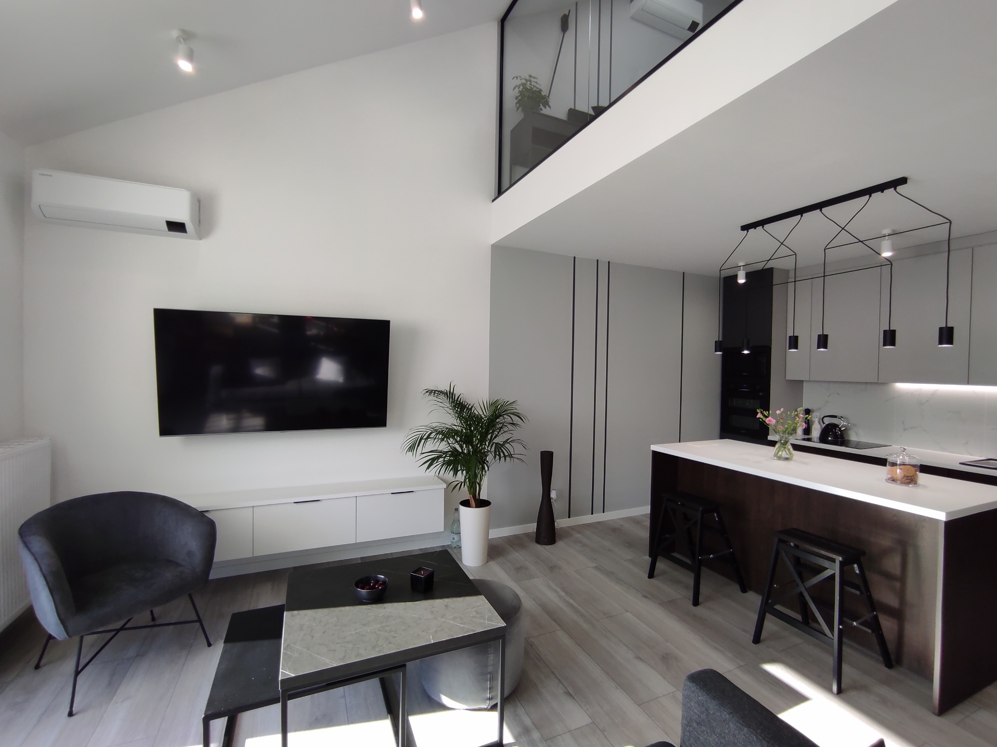 Jasne, przestronne wnętrze z antresolą, minimalistycznym salonem z szarymi meblami, telewizorem na ścianie i otwartą kuchnią z wyspą oraz designerskim oświetleniem.