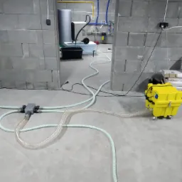 Żółty osuszacz kondensacyjny na kółkach podłączony do rozdzielacza i węży w surowym pomieszczeniu z betonowych bloczków.