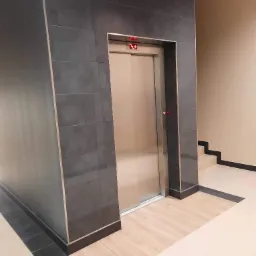 Nowoczesny hol z windą obłożoną ciemnoszarą, poziomą glazurą z metalowymi listwami, kontrastująca z jasną podłogą z płytek imitujących drewno i beżowymi ścianami.