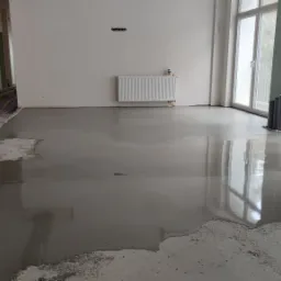 Wylewka samopoziomująca na podłodze w trakcie realizacji, widoczne kable i rury, grzejnik na ścianie, okna z widokiem na zewnątrz.