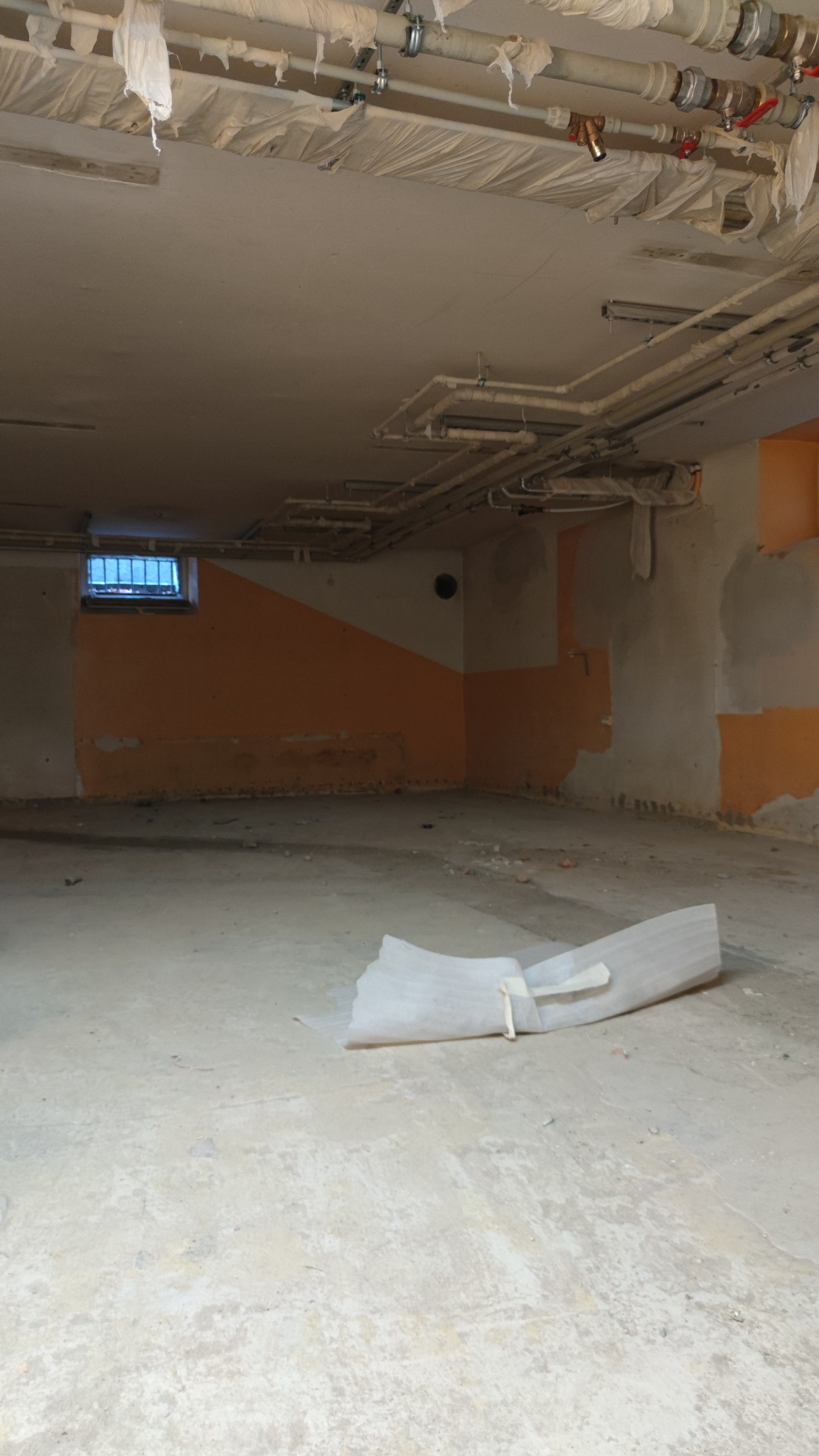 Pomieszczenie w trakcie remontu: widoczne rury pod sufitem zabezpieczone folią, fragment okna z kratą, ściany częściowo pomalowane na pomarańczowo, surowa podłoga i zwinięta folia ochronna...