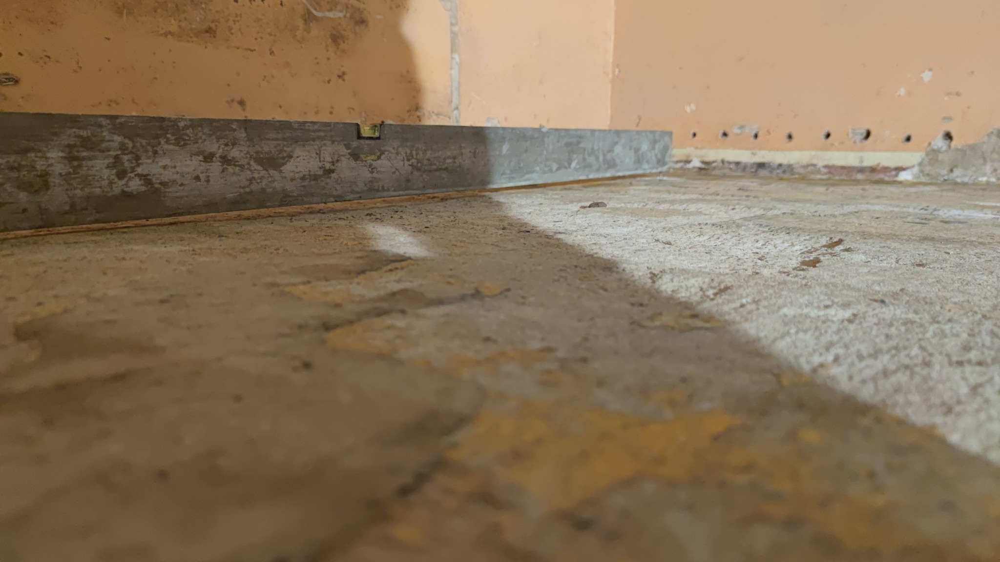 Poziomica przy ścianie na nierównym podłożu przed wylewką, widoczne ślady po starej farbie na ścianie i podłodze.