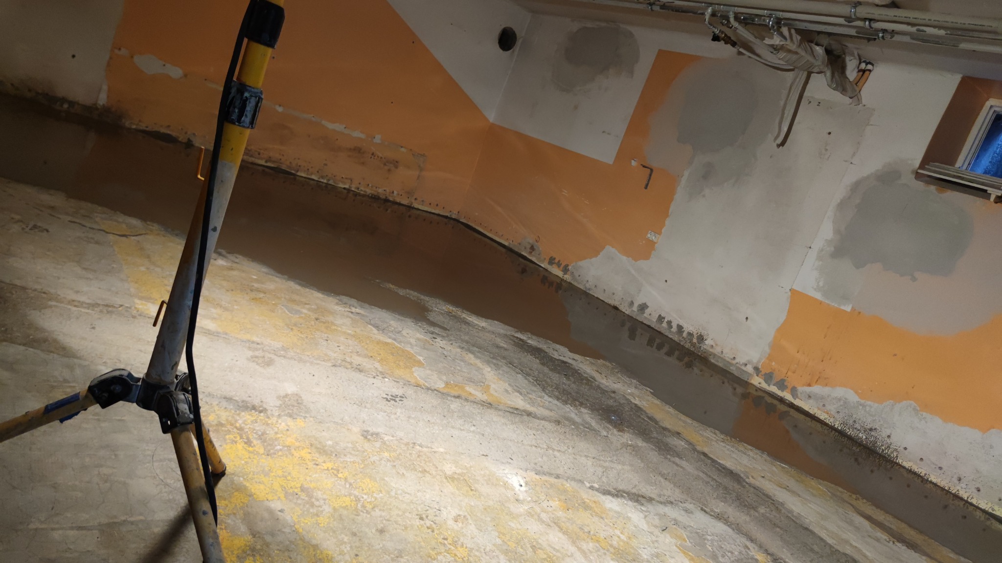 Piwnica w trakcie prac remontowych: częściowo pomalowane ściany na pomarańczowo i szaro, mokra podłoga, widoczny statyw oświetleniowy.