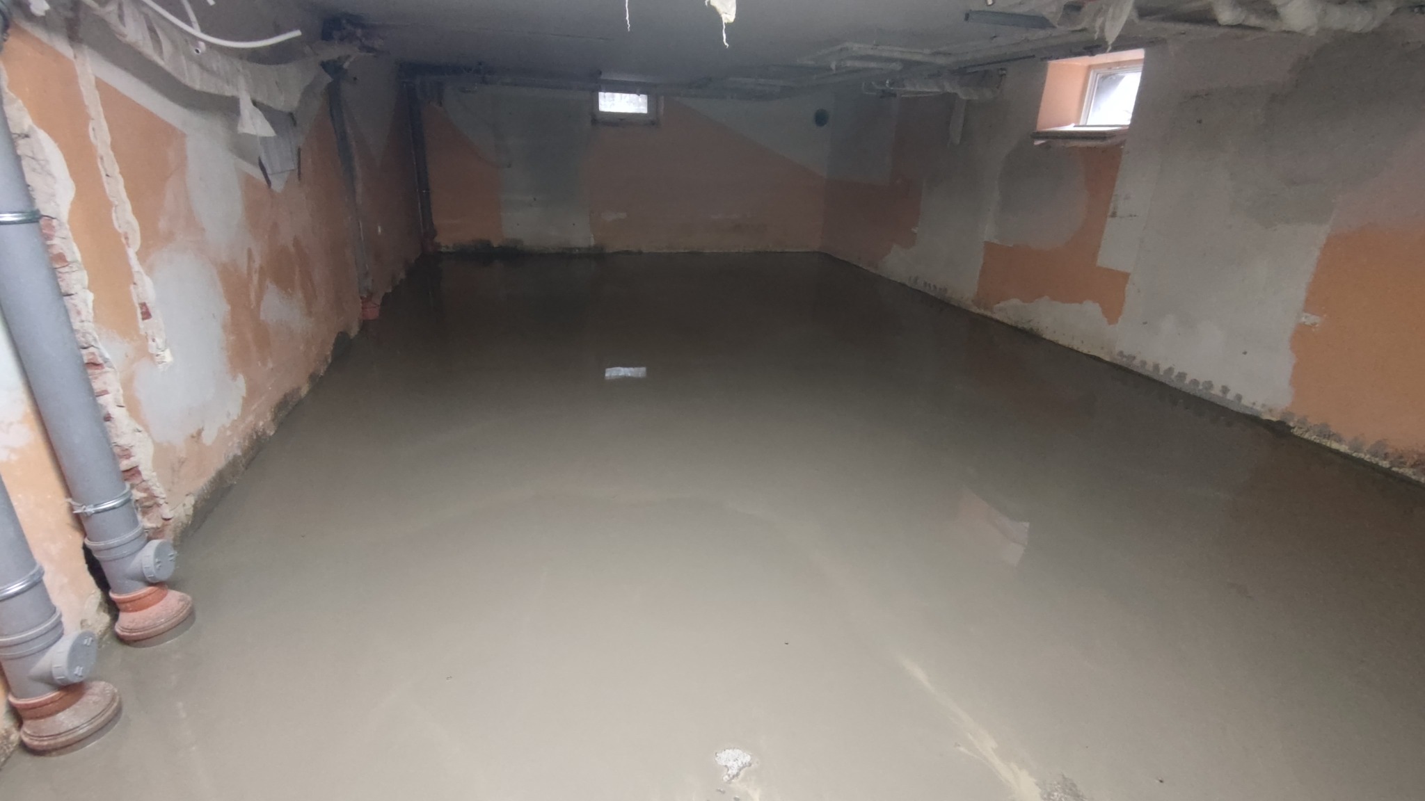 Wylewka samopoziomująca w surowym pomieszczeniu z widocznymi rurami kanalizacyjnymi przy ścianie i oknami w górnej części ścian.