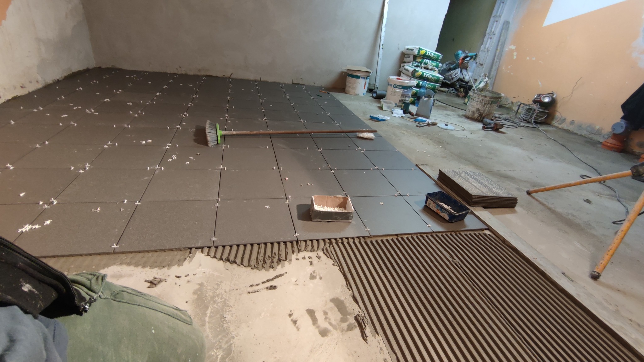 Układanie ciemnoszarych płytek podłogowych w pomieszczeniu, widoczne krzyżyki dystansowe, paczka z fugą, pędzel do rozprowadzania, narzędzia i worki z zaprawą w tle.