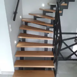 Nowoczesne schody z drewnianymi stopniami i czarną, geometryczną balustradą, oświetlone punktowymi lampkami w ścianie.