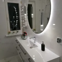 Nowoczesna łazienka z owalnym lustrem podświetlanym LED, białą umywalką z szafką, grzejnikiem drabinkowym i marmurowymi płytkami.