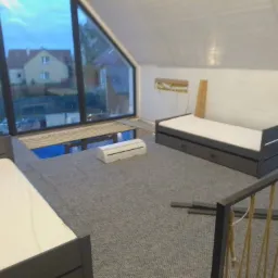 Poddasze z oknem dachowym i widokiem na domy, szara wykładzina, klimatyzator pod oknem, łóżka z szarymi ramami, częściowo wykończone barierki schodów.