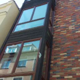 Wysoka, wąska dobudówka z oknami na ceglanej ścianie budynku w Ornecie, z widocznym odbiciem sąsiedniego budynku w szybach.