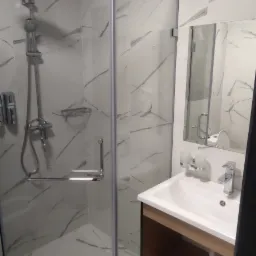 Nowoczesna łazienka z kabiną prysznicową z chromowanymi elementami, umywalką z szafką w kolorze drewna i białymi płytkami imitującymi marmur na ścianach i podłodze.