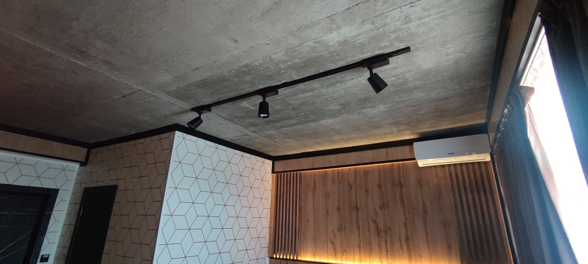 Surowy betonowy sufit z zamontowanym czarnym szynoprzewodem z trzema reflektorami, ściana z geometrycznym wzorem i drewniana ściana z podświetleniem LED, zamontowany klimatyzator.