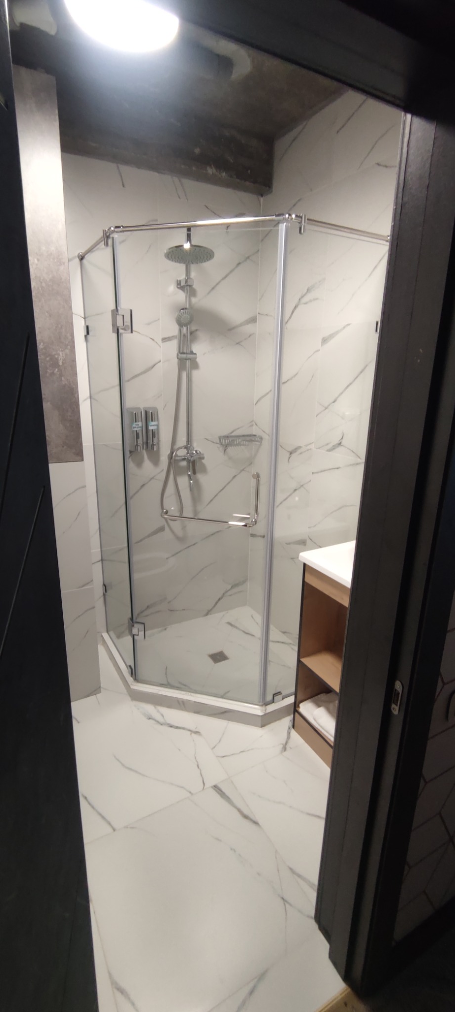 Nowoczesna kabina prysznicowa z chromowaną armaturą i szklanymi ściankami na białych płytkach imitujących marmur, widok zza uchylonych drzwi.