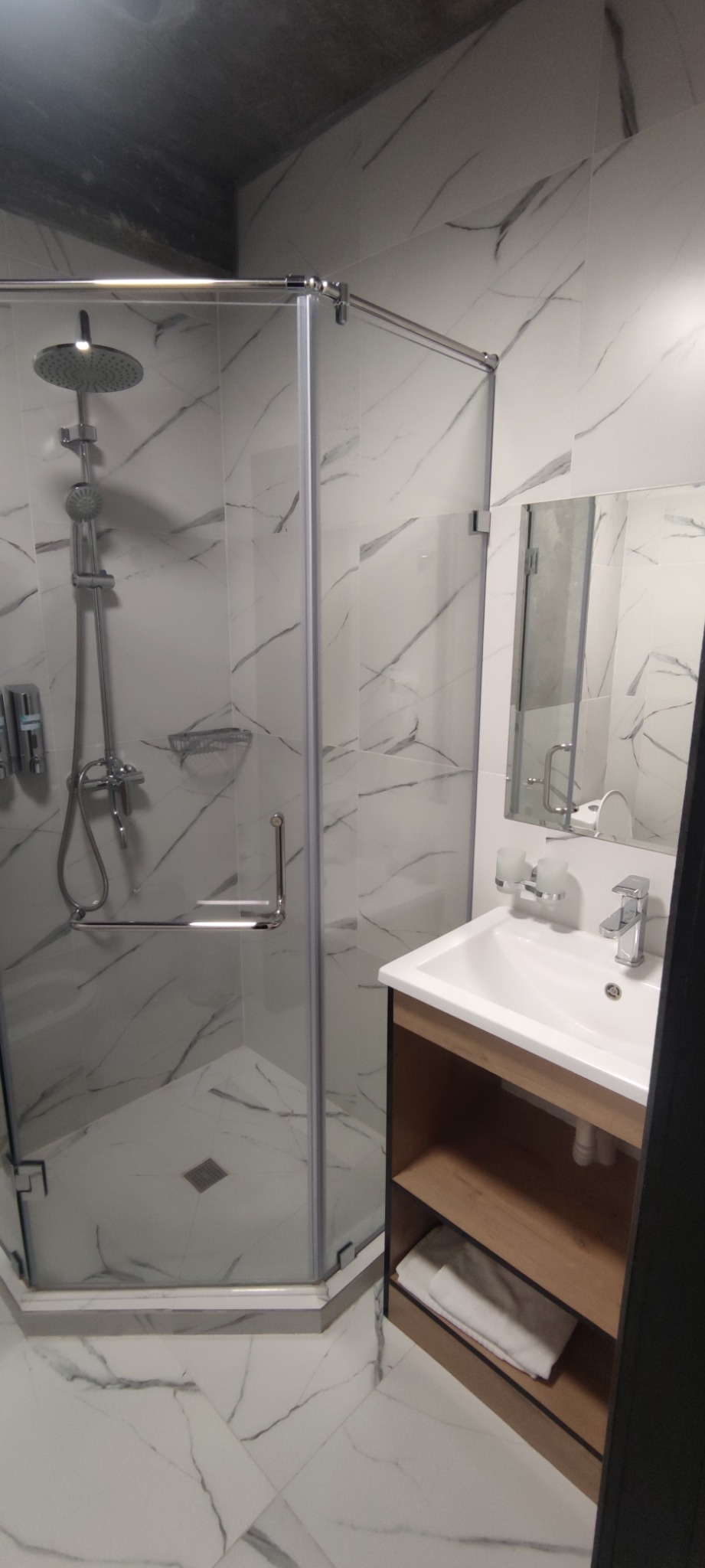 Nowoczesna łazienka z kabiną prysznicową z chromowanymi elementami, umywalką z szafką w kolorze drewna i białymi płytkami imitującymi marmur na ścianach i podłodze.