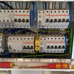 Otwarta skrzynka elektryczna z zainstalowanymi bezpiecznikami, przewodami i listwami zaciskowymi, widoczne ślady pianki montażowej na ścianie.