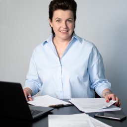 MM Finanse Monika Domagała - Kobieta w błękitnej koszuli siedzi za biurkiem z laptopem i dokumentami, uśmiechając się do kamery.