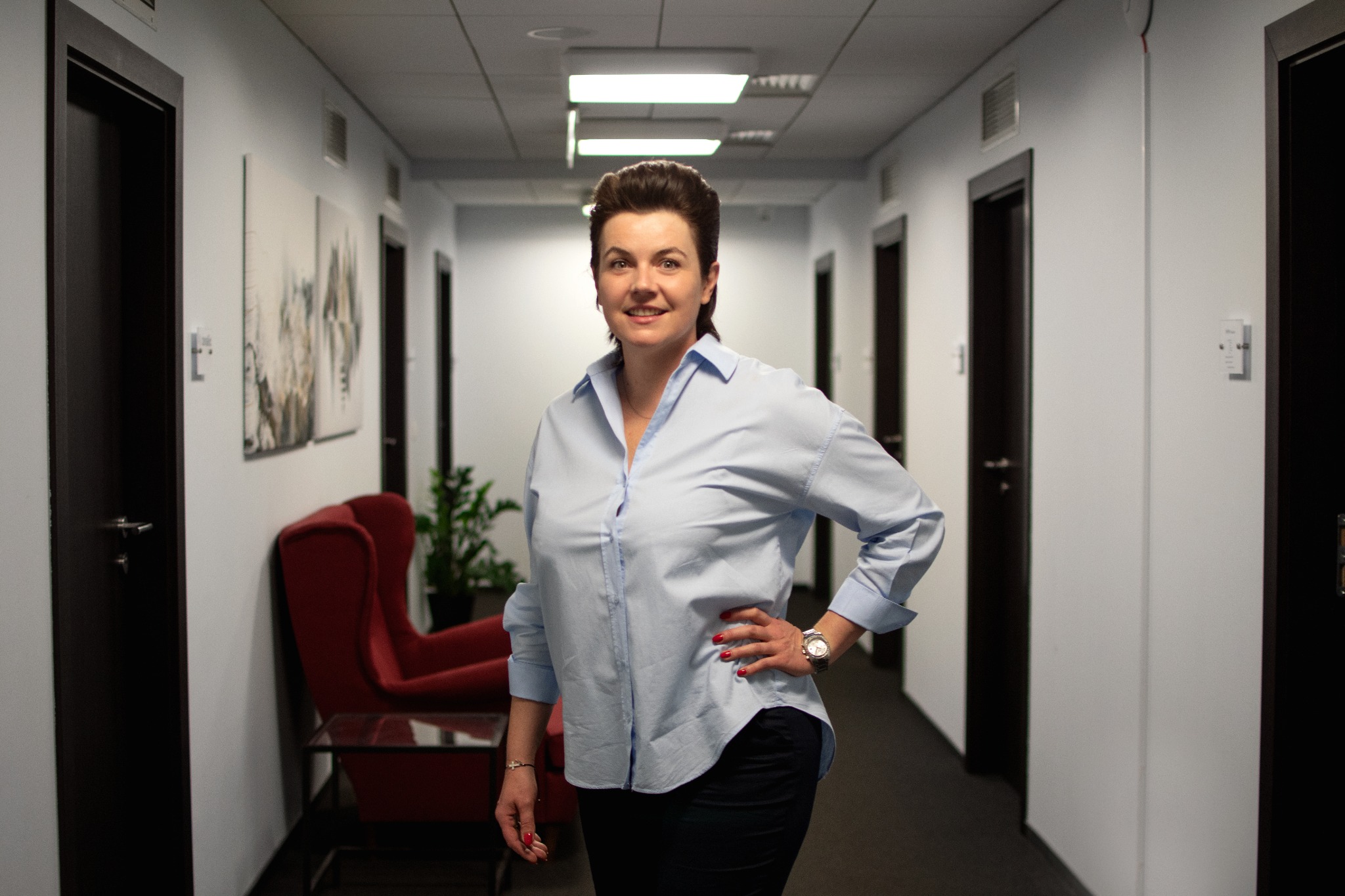 Uśmiechnięta kobieta w błękitnej koszuli stoi w jasnym korytarzu biurowym z ciemnymi drzwiami i czerwonym fotelem w tle.
