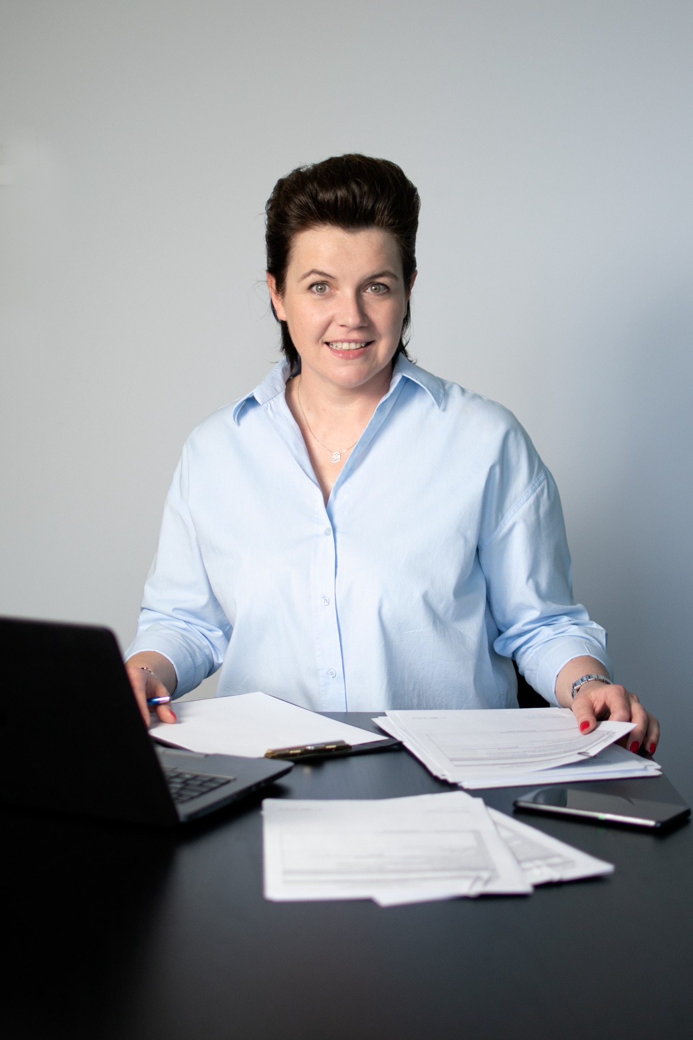 Kobieta w błękitnej koszuli siedzi za biurkiem z laptopem i dokumentami, uśmiechając się do kamery.