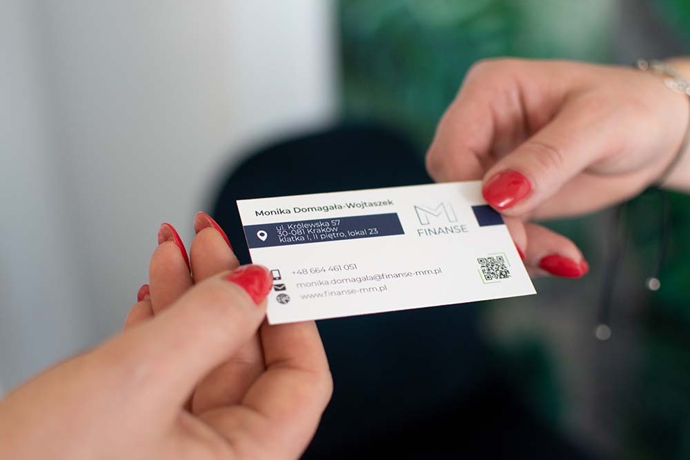 Dwie dłonie wymieniają się wizytówką firmy finansowej, z adresem w Krakowie, numerem telefonu i adresem strony internetowej, paznokcie pomalowane na czerwono.