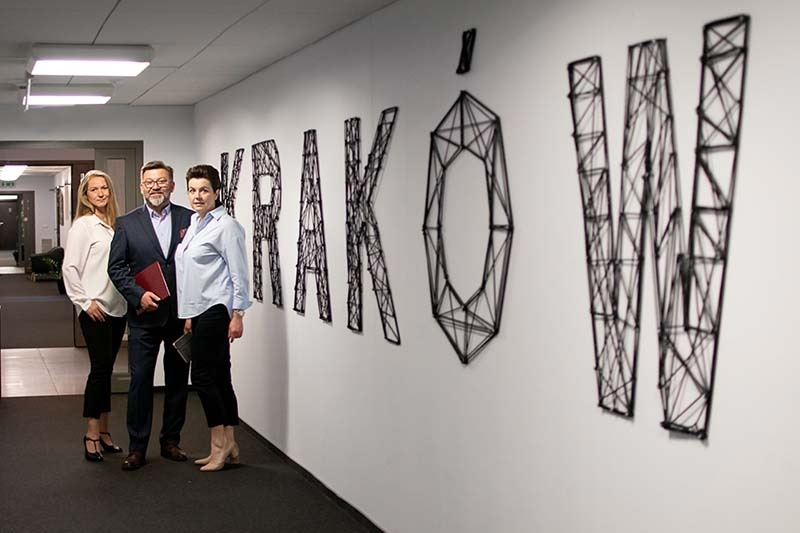 Trzy osoby stojące w korytarzu biurowym na tle ściany z napisem 'KRAKÓW' wykonanym z czarnego sznurka.