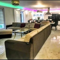 Remonty i usługi budowlane Sebastian Jacak - Nowoczesny salon z narożną sofą w odcieniach brązu i beżu, marmurową podłogą, oświetleniem LED i widokiem na otwartą kuchnię.