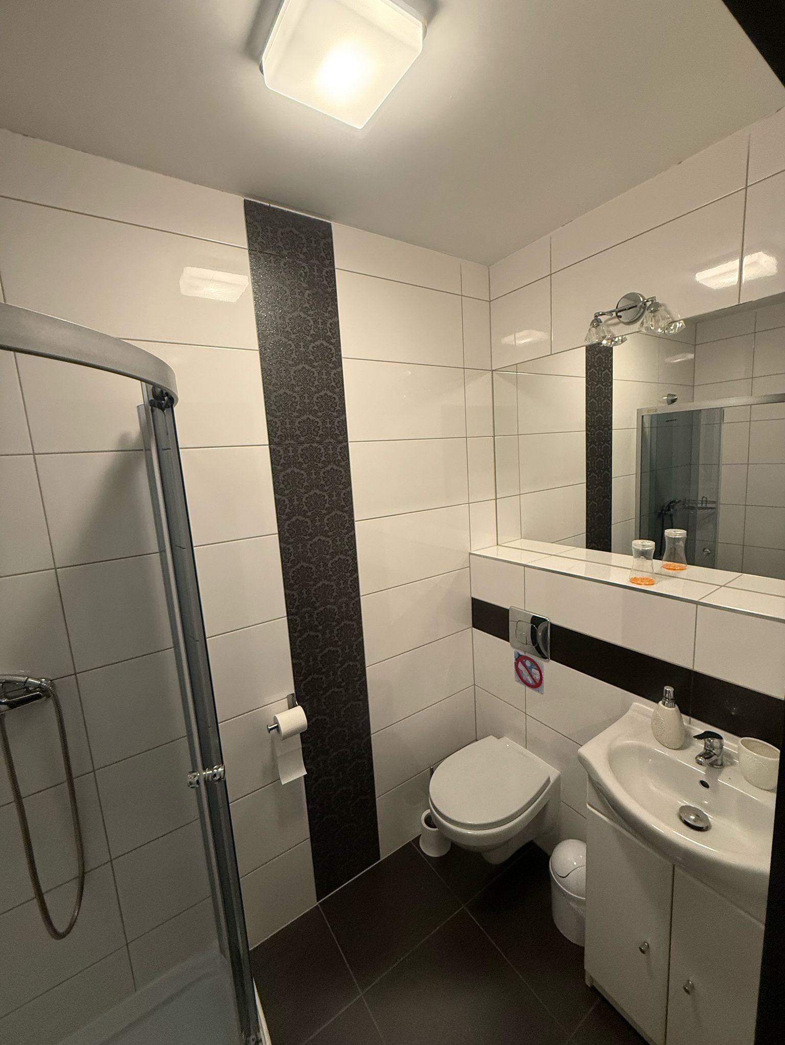 Nowoczesna łazienka z kabiną prysznicową, białą ceramiką i akcentem w postaci pionowego pasa płytek w kolorze ciemnego grafitu z ornamentem, oświetlona kwadratową lampą sufitową.