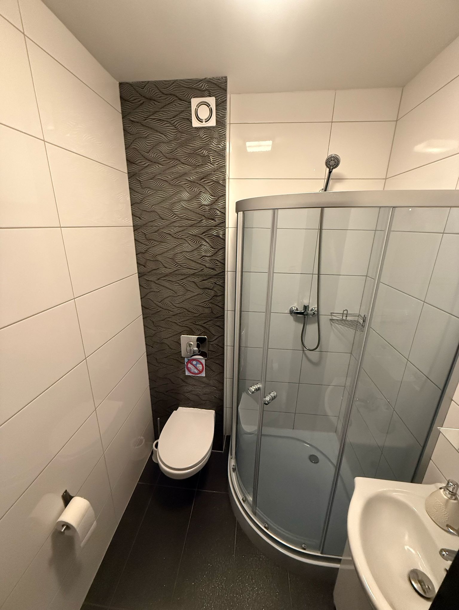 Nowoczesna łazienka z czarną podłogą, białą armaturą i kabiną prysznicową z zaokrąglonymi drzwiami, ściana z dekoracyjną, falistą fakturą płytek.