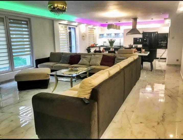 Nowoczesny salon z narożną sofą w odcieniach brązu i beżu, marmurową podłogą, oświetleniem LED i widokiem na otwartą kuchnię.