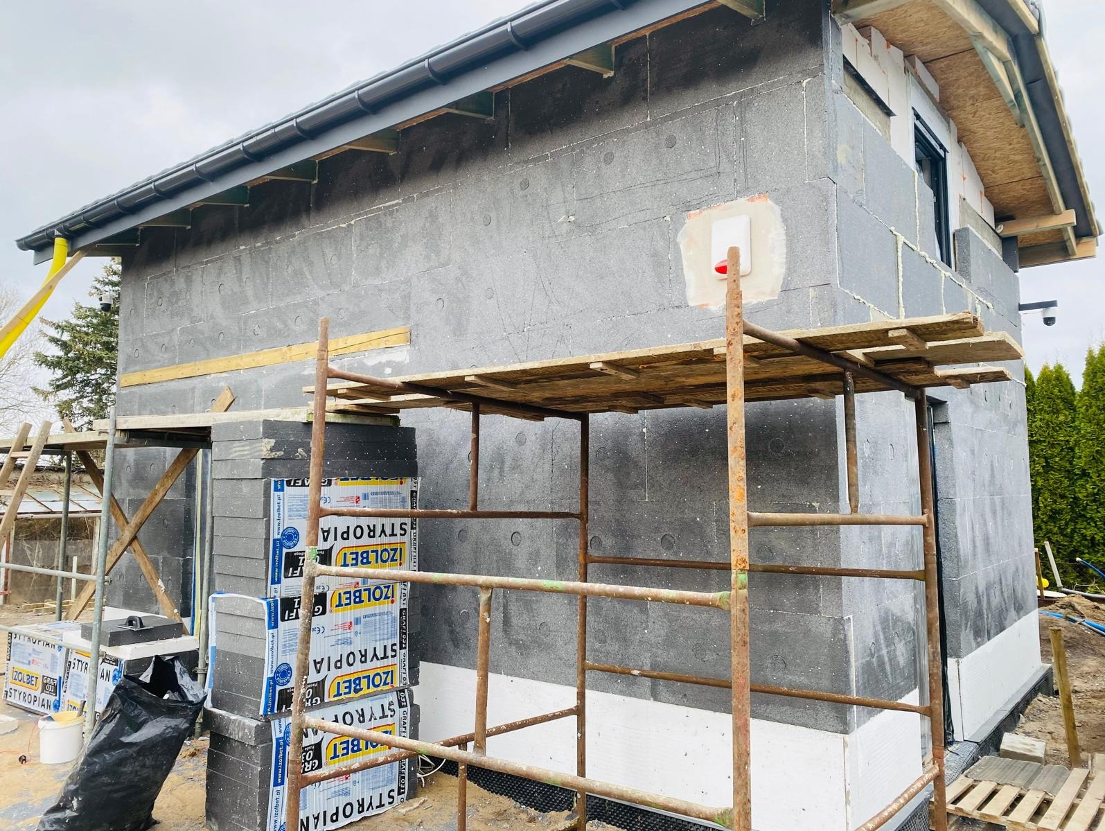 Ocieplanie elewacji domu styropianem grafitowym, widoczne rusztowanie, paczki styropianu Izolet i fragment elewacji z zamontowanym ociepleniem oraz brakujące miejsce po koszu do koszykówki.
