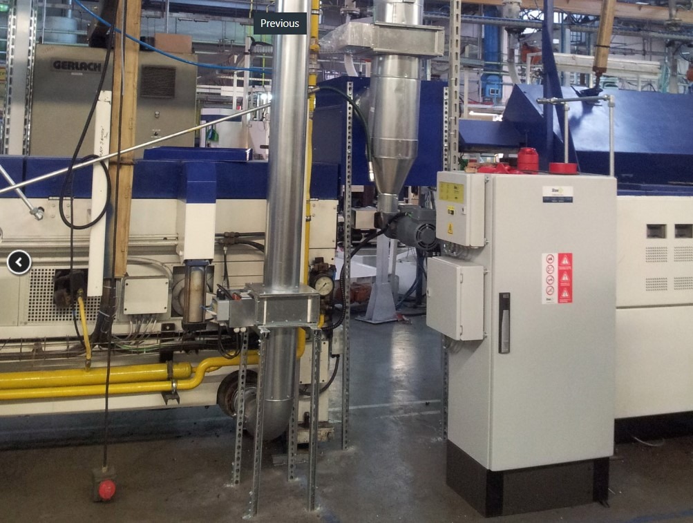 Widok instalacji przemysłowej z rurami wentylacyjnymi, skrzynką sterowniczą z ostrzeżeniami i żółtymi rurami gazowymi w tle hali produkcyjnej.