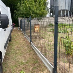 Vasylenko Viacheslav - Nowo zainstalowane, panelowe ogrodzenie z podmurówką betonową, widoczne z boku, z pracownikiem w tle i białym vanem po lewej stronie, na tle zieleni i silosów zbożowych.