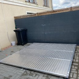 Vasylenko Viacheslav - Metalowa platforma z antypoślizgową fakturą, prawdopodobnie element antresoli, leżąca na brukowanej powierzchni przed czarnym ekranem i ścianą budynku.