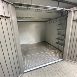 Vasylenko Viacheslav - Wnętrze otwartego, metalowego schowka ogrodowego z podłogą w romby i półkami po prawej stronie, stojącego na kostce brukowej.