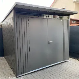 Nowoczesny, metalowy schowek ogrodowy z dwuskrzydłowymi drzwiami, umieszczony na kostce brukowej, z widocznym fragmentem domu w tle.